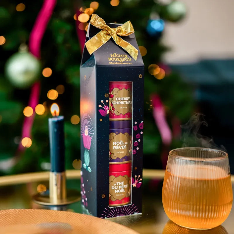 Coffret Trilogie enchantée Maison Bourgeon avec ses trois boîtes, un ruban doré et une tasse fumante devant un sapin de Noël.