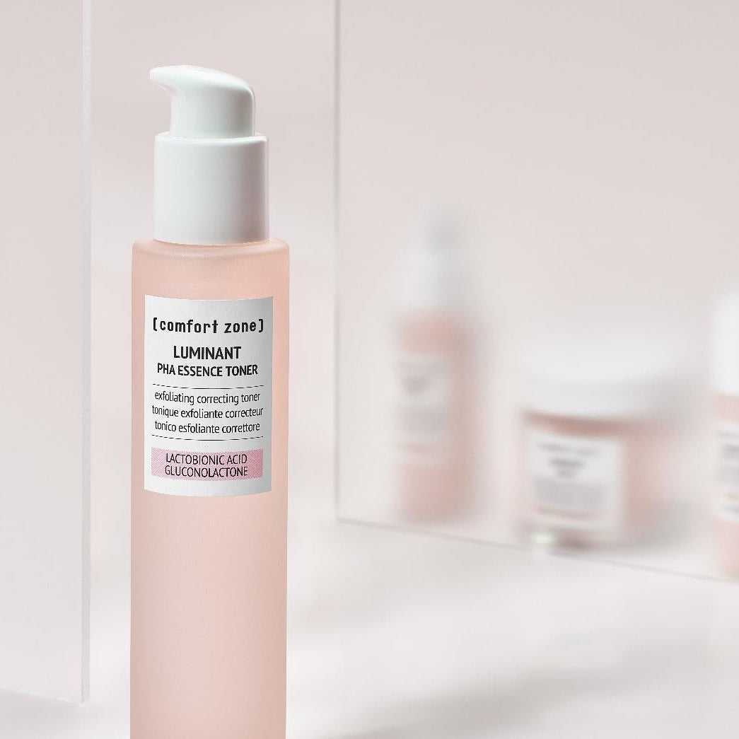 Flacon rose Luminant PHA Essence Toner de Comfort Zone, devant un miroir reflétant les autres soins de la gamme.