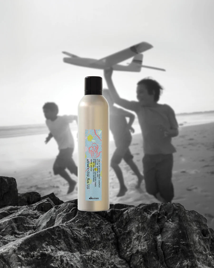 Laque Davines This Is An Extra Strong Hair Spray 400 ml posée sur un rocher, devant une photographie en noir et blanc d’enfants jouant sur la plage.
