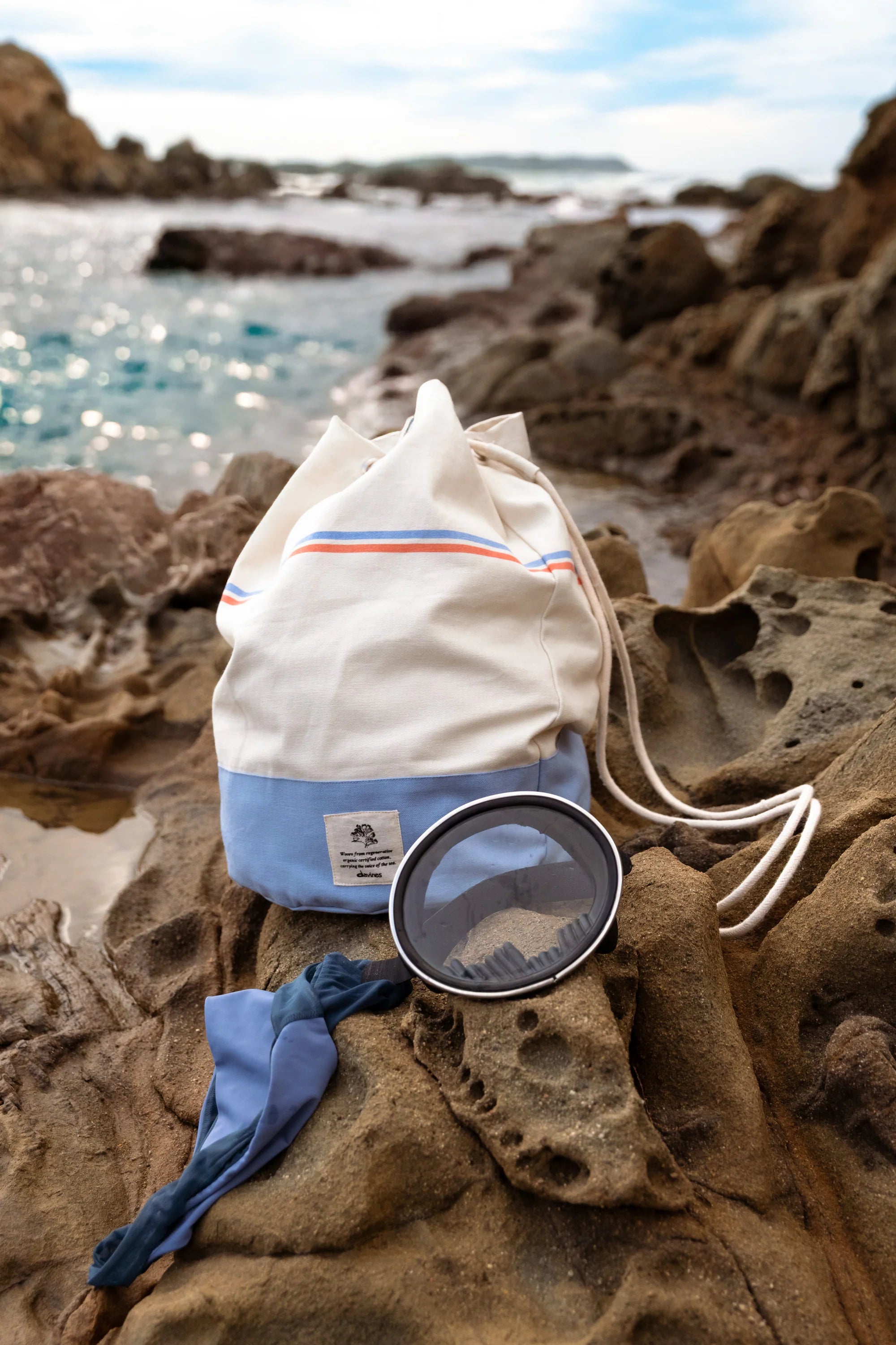 Sac à dos Davines SU en coton posé sur les rochers en bord de mer, accessoire estival pour transporter ses essentiels cheveux et corps après l’exposition solaire