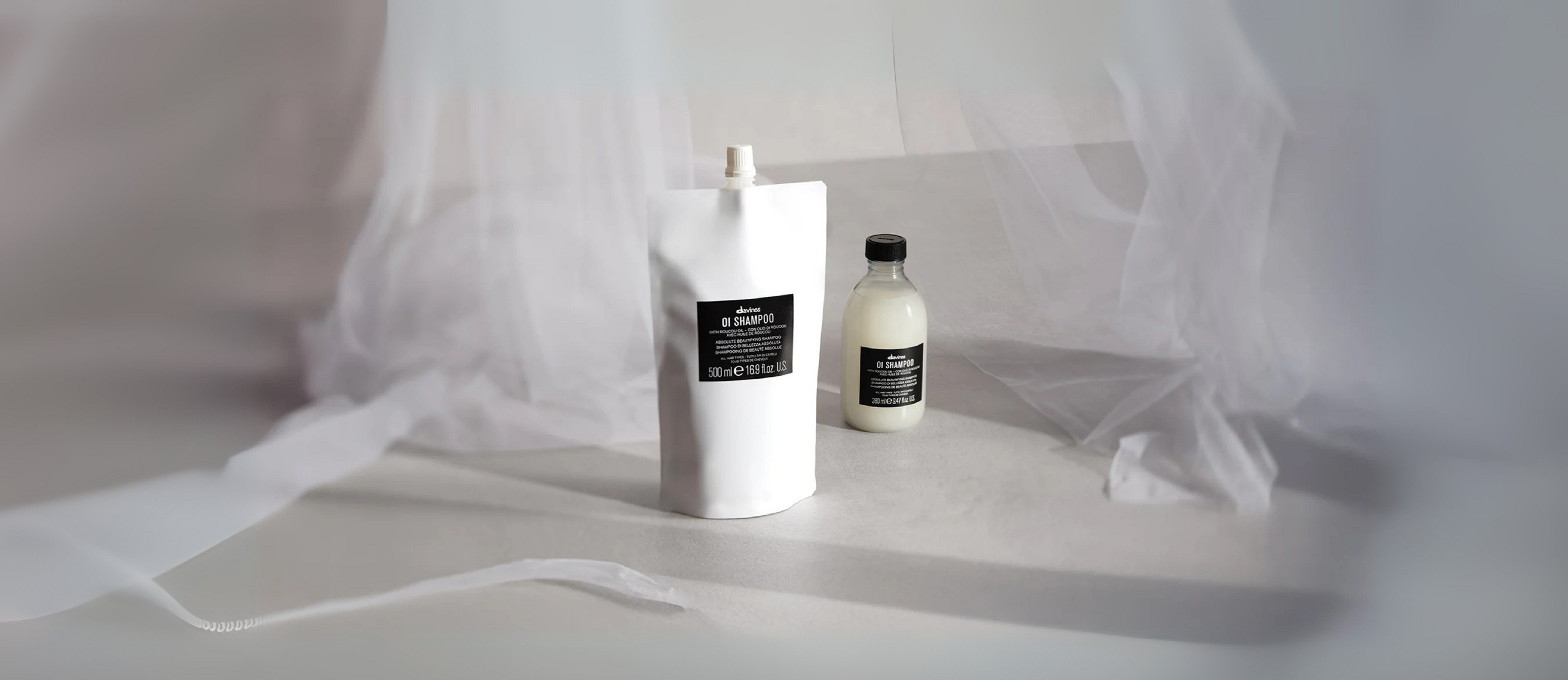 Éco-recharge Davines OI Shampoo 500 ml posée à côté du flacon verre 280 ml, sur fond neutre voilé de tissu léger.