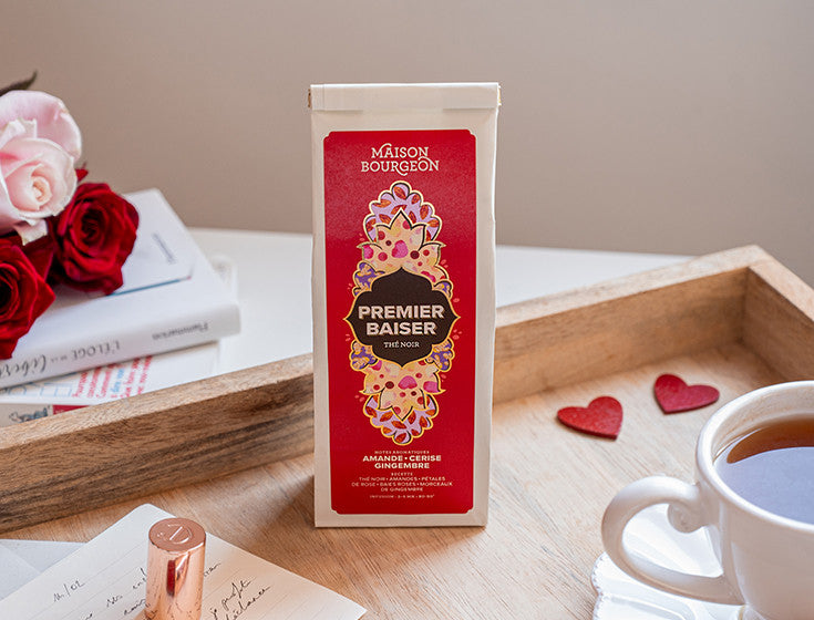 Paquet de thé noir Maison Bourgeon Premier Baiser avec des illustrations de roses en arrière-plan, posé sur un plateau en bois avec une tasse de thé.