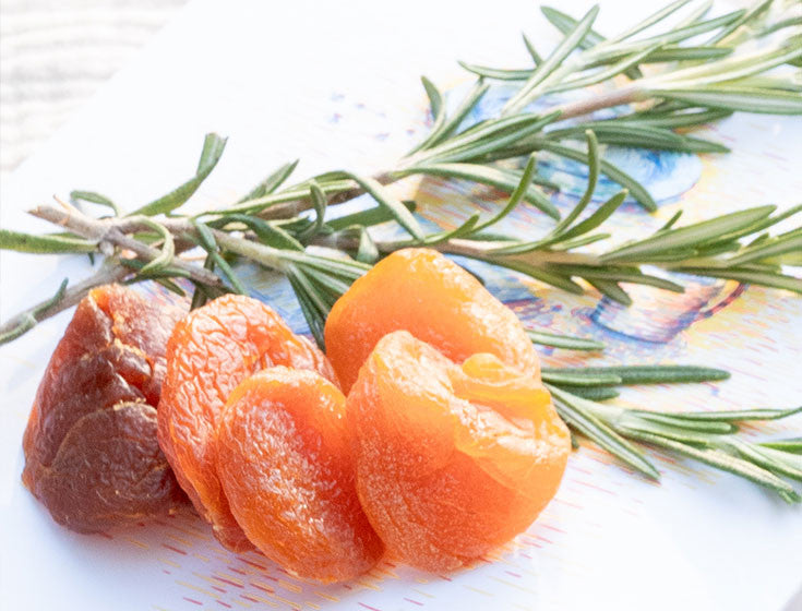 Gros plan sur des abricots secs et une branche de romarin sur une assiette blanche.