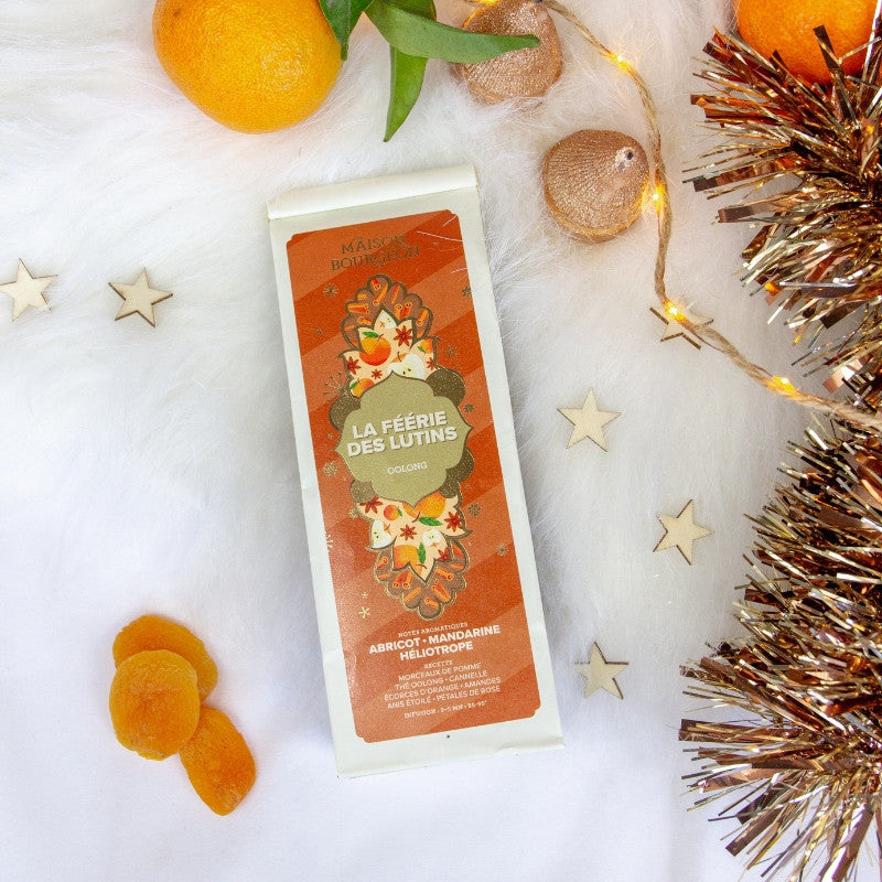 Thé Oolong La Féérie des Lutins de Maison Bourgeon en ambiance festive de Noël. Mélange aromatisé aux notes d'abricot, mandarine et héliotrope. Infusion hivernale et gourmande, idéale pour les fêtes, avec du thé Oolong, morceaux de pomme, cannelle, écorces d'orange, amandes, anis étoilé et pétales de rose. Présenté sur un décor cosy avec des agrumes, des étoiles et des guirlandes dorées.