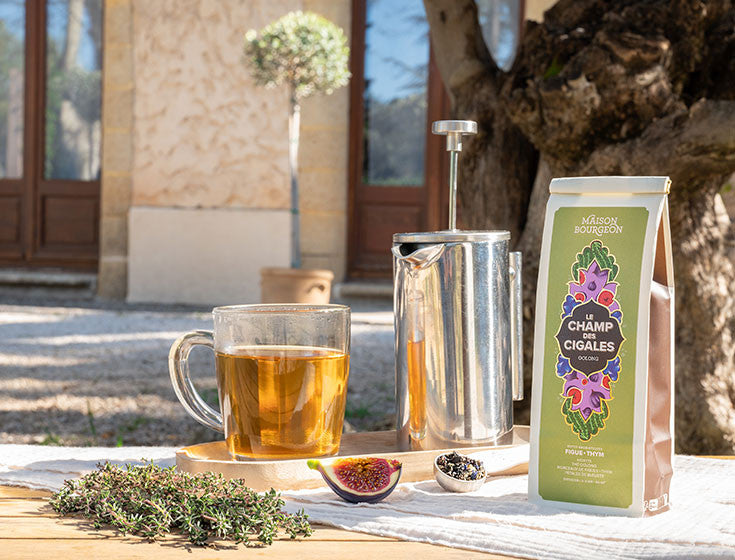 Thé Oolong Le Champ des Cigales de Maison Bourgeon présenté en extérieur, avec une théière en métal, une tasse de thé, des branches de thym frais, une figue coupée et une petite cuillère de feuilles de thé. L'image évoque une dégustation de thé dans un cadre naturel.