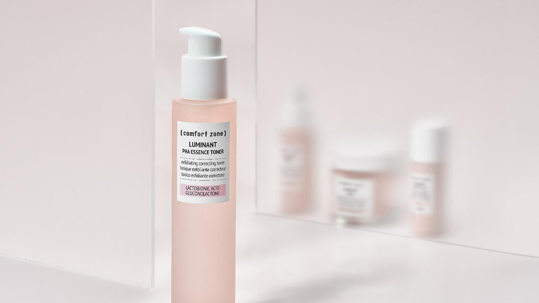 Flacon rose Luminant PHA Essence Toner de Comfort Zone, devant un miroir reflétant les autres soins de la gamme.
