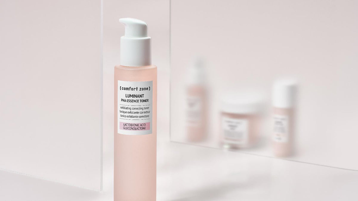 Flacon rose Luminant PHA Essence Toner de Comfort Zone, devant un miroir reflétant les autres soins de la gamme.