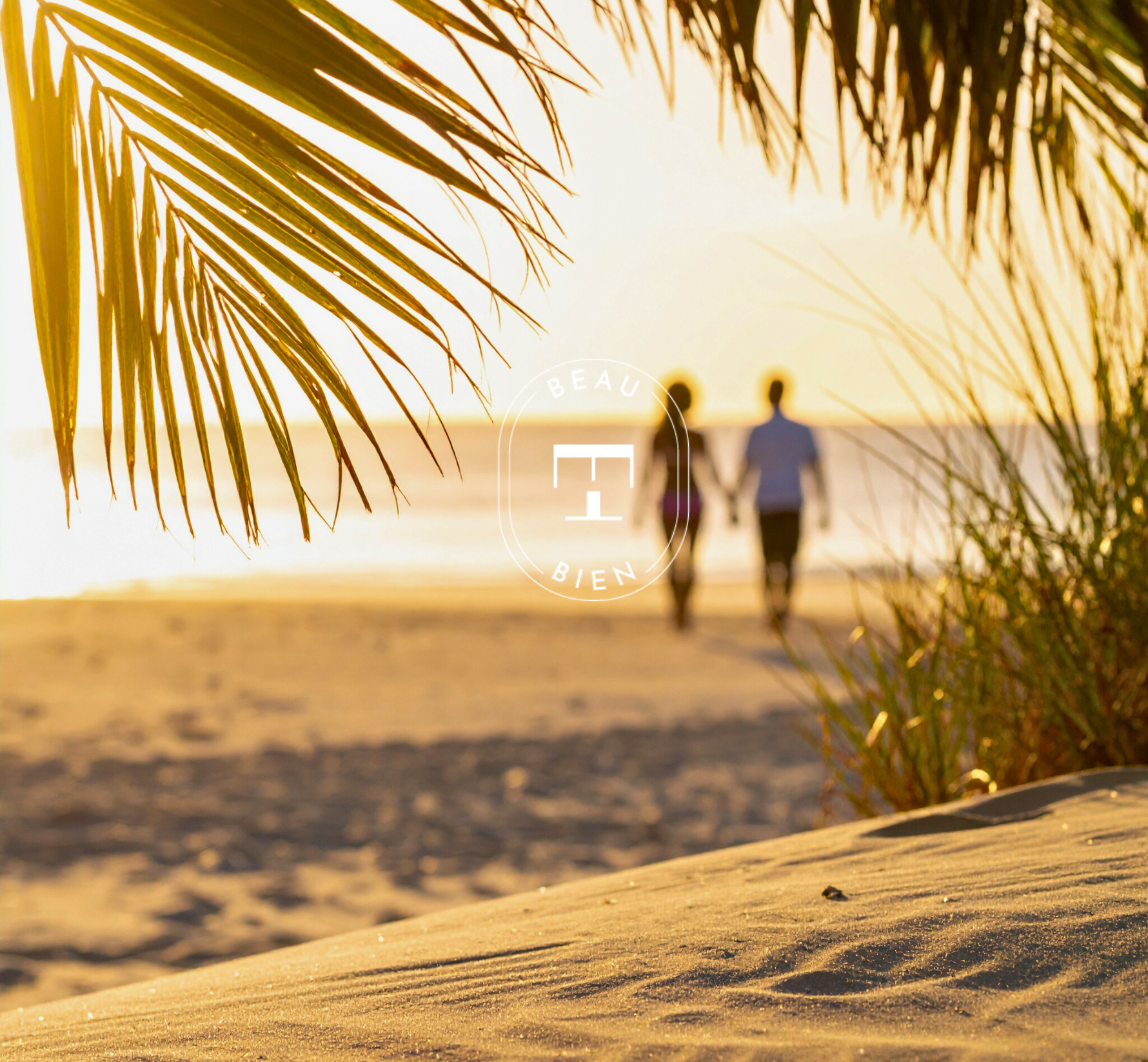 Silhouettes d’un homme et d’une femme marchant main dans la main sur une plage au coucher du soleil, encadrées par des herbes de dune et des feuilles de palmier au premier plan.