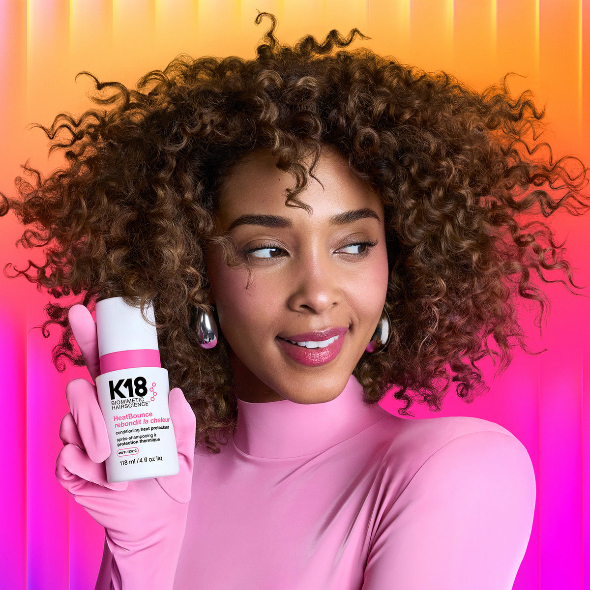 Femme aux cheveux bruns bouclés, vêtue de rose, tenant un flacon K18 HeatBounce près de son visage sur fond rose et orange.