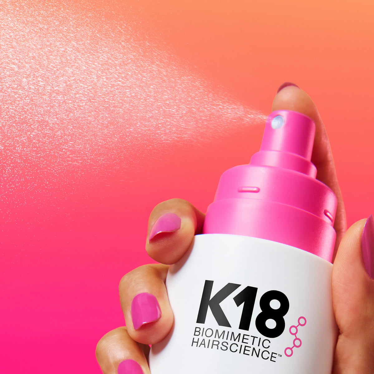 Main aux ongles roses actionnant le vaporisateur K18 HeatBounce, qui diffuse une fine brume sur fond rose et orange.