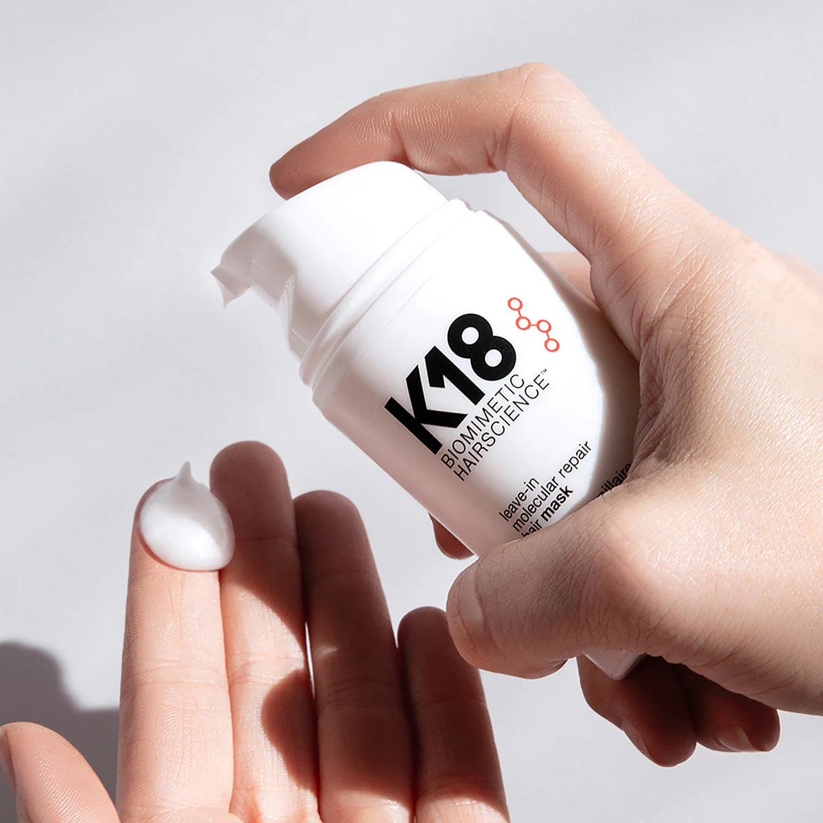 Une main tient le flacon-pompe du masque K18 au-dessus d’un doigt portant une noisette de crème blanche.