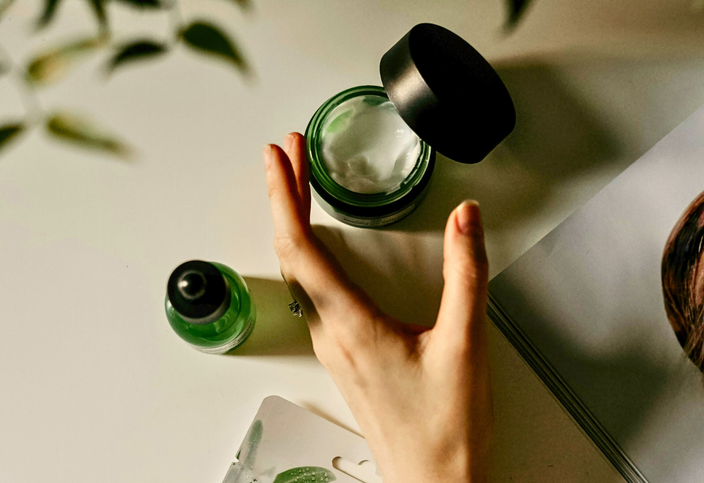 Une main applique de la crème hydratante, posée sur une table avec un flacon de sérum vert et une feuille verte en arrière-plan, représentant le soin et le contact avec la nature.