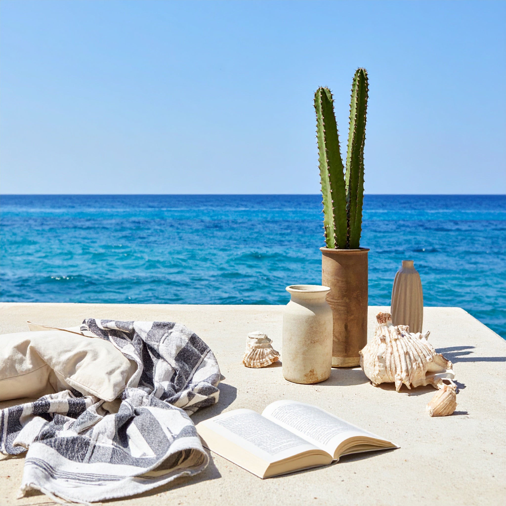 Plage méditerranéenne minimaliste : table en pierre face à la mer bleu azur, cactus en pot, coquillages, coussin, plaid rayé et livre ouvert sous une lumière douce d’été.







