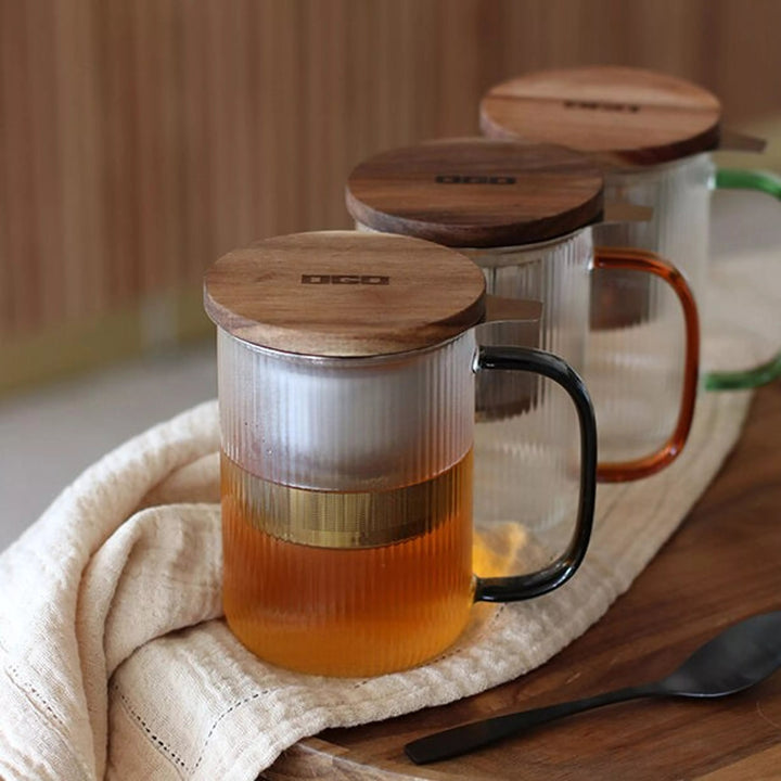 Trois tisanieres en verre borosilicate strie de 475 ml avec couvercles bois, filtre inox et anses colorees (noire, ambree, verte), de la marque Ogo Living, presentees sur un plateau en bois dans un cadre chaleureux.