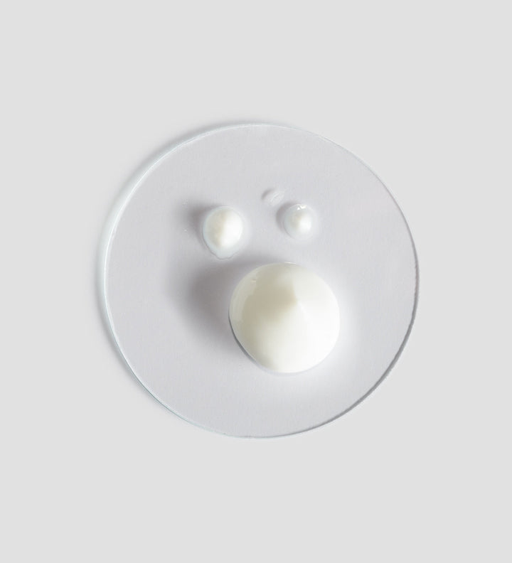 Texture du Comfort Zone Essential Peeling, trois gouttes de crème blanche sur une surface circulaire transparente, sur fond gris clair.