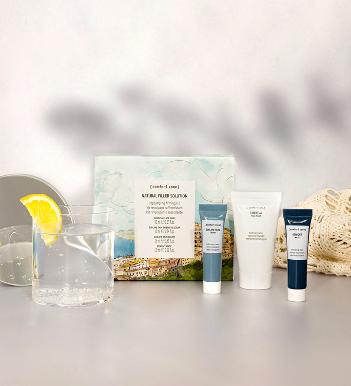 Un ensemble de produits de soin de la marque Comfort Zone intitulé "Natural Filler Solution". L'ensemble inclut plusieurs tubes de produits placés devant une boîte d'emballage illustrée. À côté des produits, un verre d'eau avec une tranche de citron ajoute une touche de fraîcheur. L'image présente une ambiance lumineuse et propre, avec un fond blanc et quelques accessoires de soin. Cette composition met en avant les produits de la marque, soulignant leur qualité et leur attrait pour les soins de la peau.