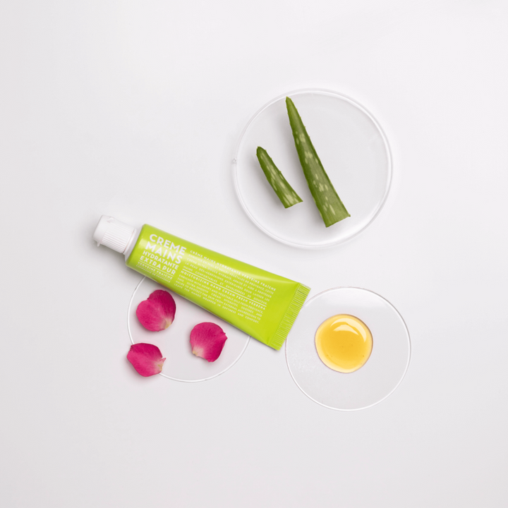 Tube de crème mains hydratante Extra Pur à la verveine fraîche de la marque Compagnie de Provence, posé sur une surface blanche. À côté, des pétales de rose, des morceaux d'aloe vera et une goutte de miel, illustrant les ingrédients naturels de la composition.