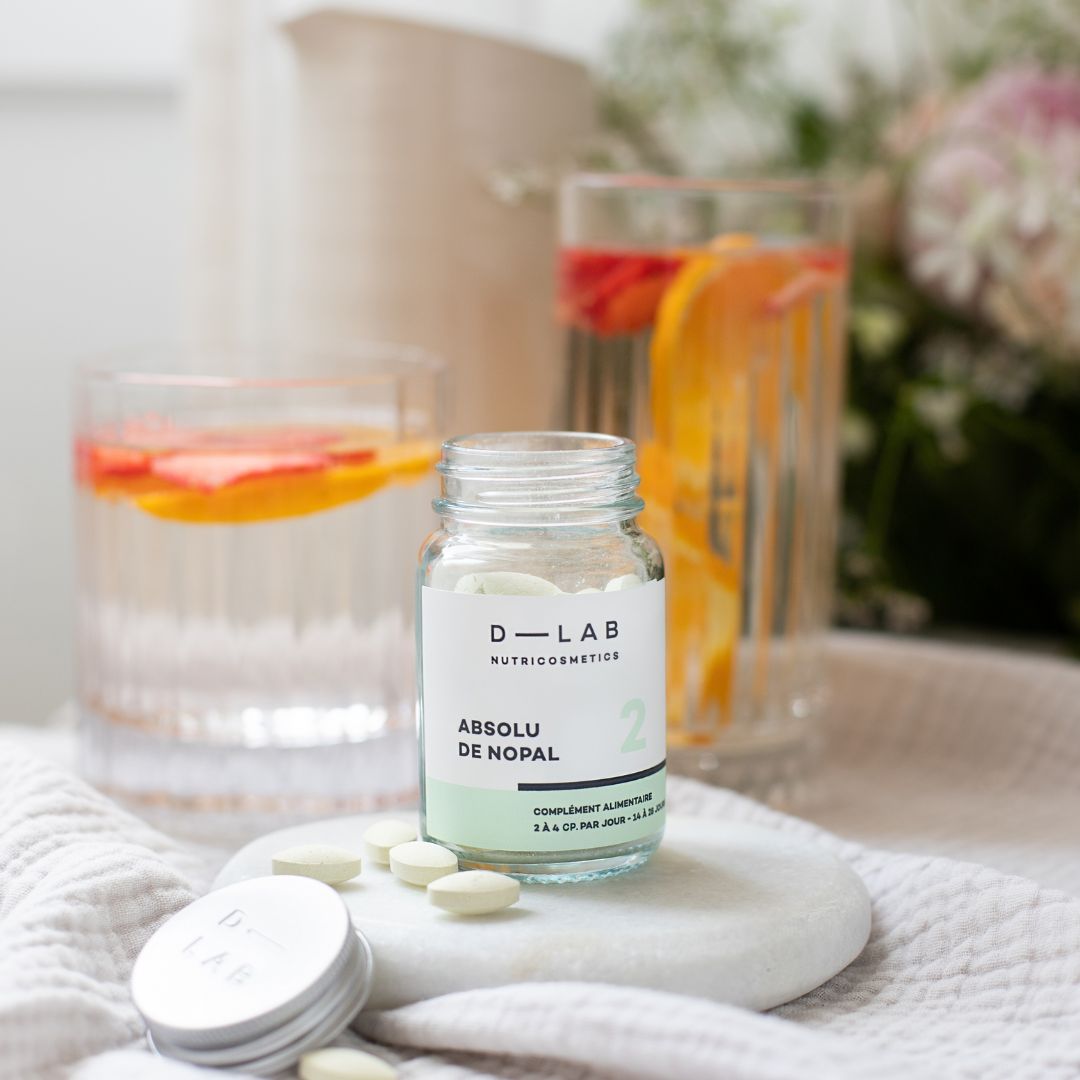 Bouteille de compléments alimentaires D-LAB Nutricosmetics Absolu de Nopal posée sur un tissu, avec des verres d'eau aromatisée en arrière-plan.