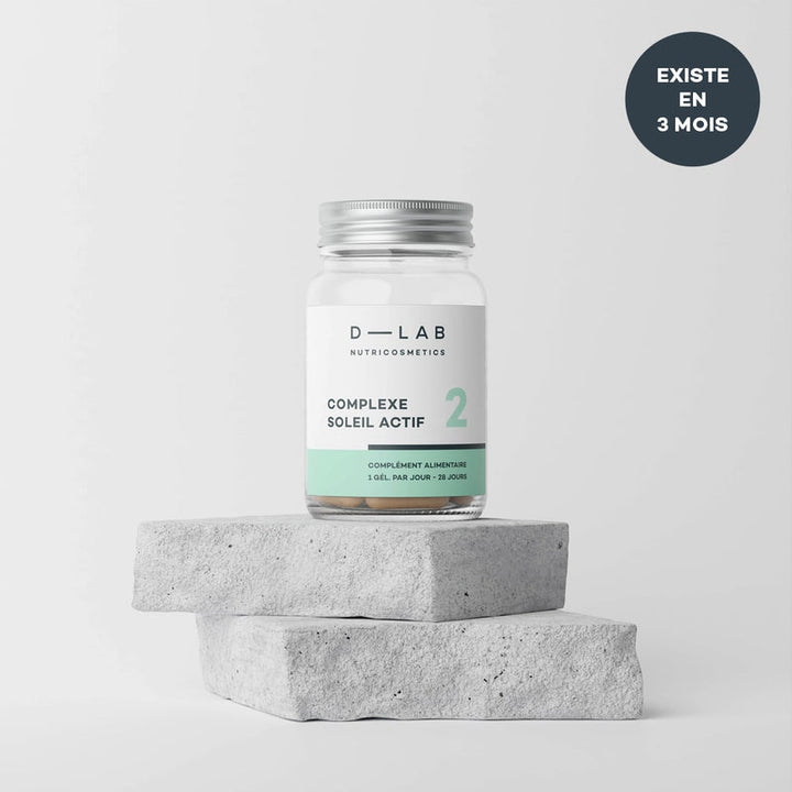 Bouteille de complément alimentaire "Complexe Soleil Actif" de D-Lab Nutricosmetics. La bouteille est placée sur deux blocs de pierre grise et présente un design épuré avec une étiquette blanche et verte. Inscription sur la bouteille : "1 gélule par jour - 28 jours". Texte dans un cercle noir en haut à droite : "Existe en 3 mois".