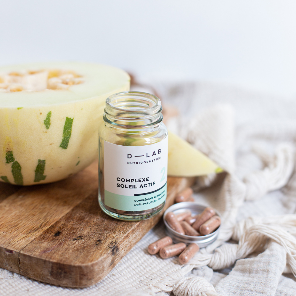 Flacon ouvert de capsules du "Complexe Soleil Actif" de D-Lab Nutricosmetics, posé sur une planche en bois à côté d'une tranche de melon. Capsules dans un petit couvercle à côté du flacon, sur un fond de tissu beige.