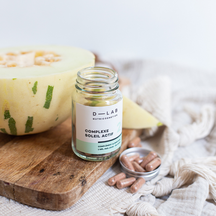 Flacon ouvert de capsules du "Complexe Soleil Actif" de D-Lab Nutricosmetics, posé sur une planche en bois à côté d'une tranche de melon. Capsules dans un petit couvercle à côté du flacon, sur un fond de tissu beige.