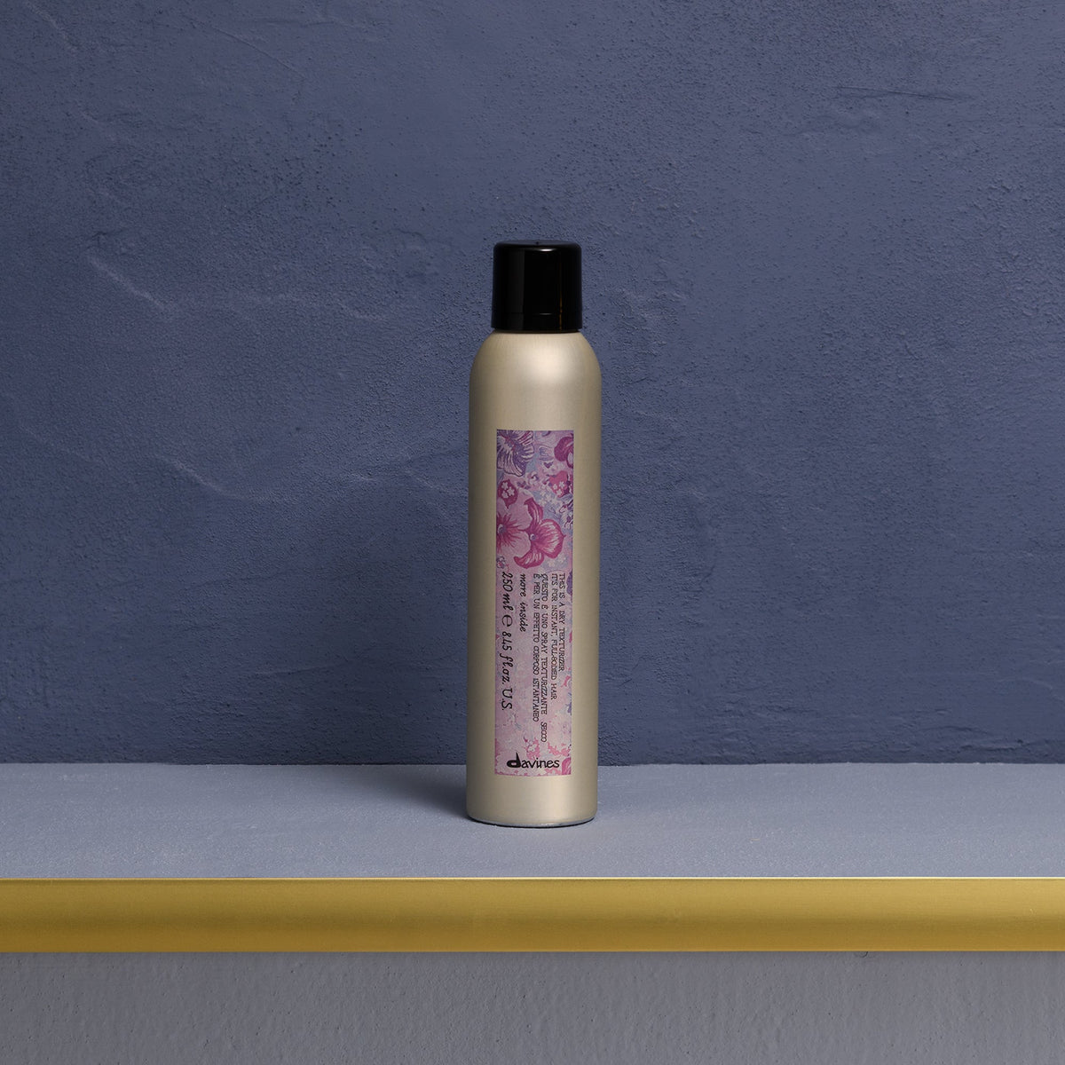 Spray texturisant Davines This Is A Dry Texturizer 250 ml posé sur une étagère dorée devant un mur bleu, avec son étiquette florale violette.