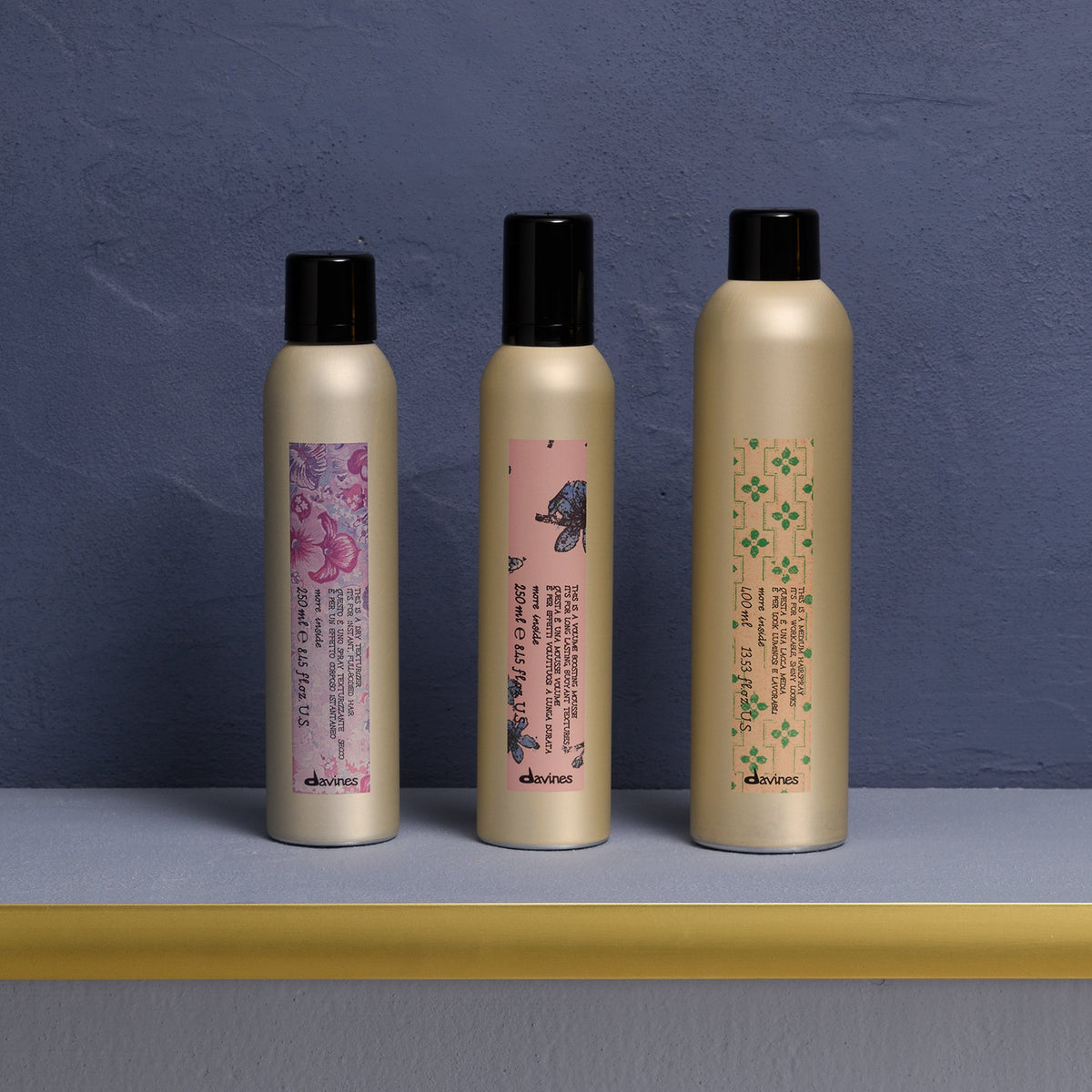 Davines Dry Texturizer 250 ml, Volume Boosting Mousse 250 ml et Medium Hairspray 100 ml réunis sur une étagère dorée devant un mur bleu ; photo de la routine More Inside.