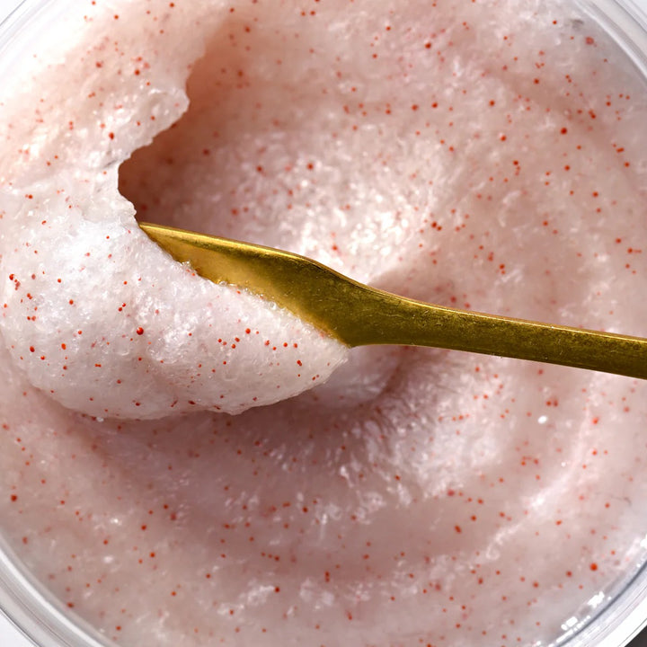 Gros plan de la texture rose cristalline du gommage SOLU, grains de sel marin visibles avec spatule dorée.