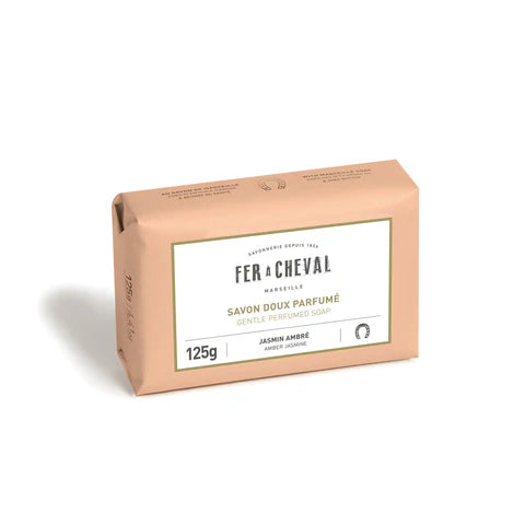 Image d'un savon doux parfumé de la marque Fer à Cheval, parfum jasmin ambré, de 125g. Le savon est emballé dans un papier de couleur pêche clair avec une étiquette blanche au centre portant le nom de la marque, le type de produit et son parfum. Le design simple et élégant reflète la qualité artisanale et naturelle du produit.