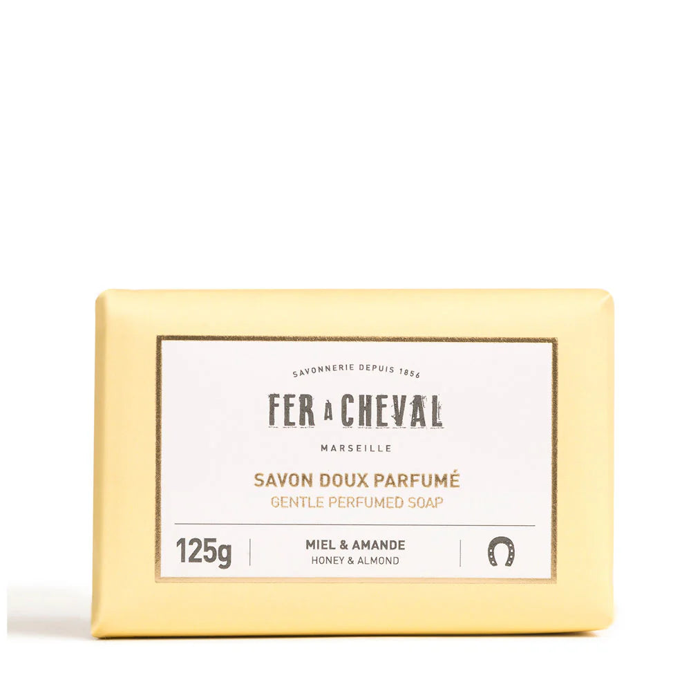 Image d'un savon doux parfumé de la marque Fer à Cheval, parfum miel et amande, de 125g. Le savon est emballé dans un papier de couleur crème avec une étiquette blanche au centre portant le nom de la marque, le type de produit et son parfum. Le design simple et élégant reflète la qualité artisanale et naturelle du produit.