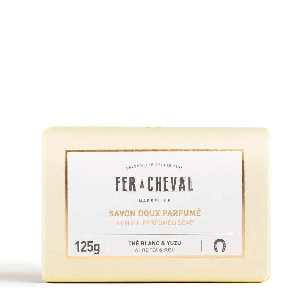 Image du savon doux parfumé de la marque Fer à Cheval, côté dos de l'emballage. Le paquet est de couleur crème avec des inscriptions en noir, incluant des informations sur les ingrédients et les instructions d'utilisation. Le savon est parfumé au thé blanc et yuzu, et on peut également voir le poids de 125g sur l'emballage.