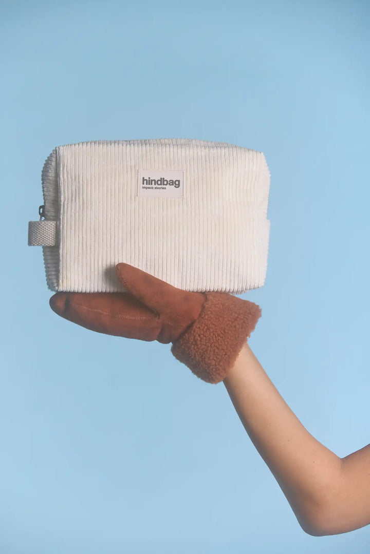 Trousse de toilette en velours côtelé écru de la marque Hindbag, tenue en équilibre sur une main gantée en cuir marron avec doublure en fausse fourrure, sur un fond bleu clair. Ambiance hivernale et minimaliste.
