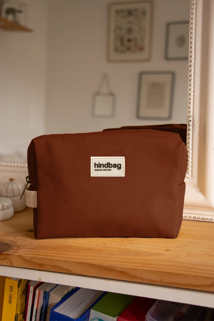 Trousse de toilette en toile de coton bio couleur marron chocolat de la marque Hindbag, posée sur une étagère en bois devant un miroir encadré de blanc. En arrière-plan, des objets de décoration et des cadres accrochés au mur, apportant une ambiance chaleureuse et minimaliste.
