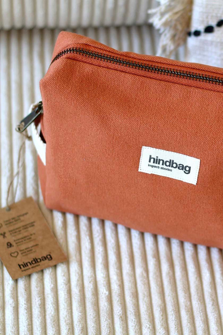 Trousse de toilette en toile de coton bio couleur terracotta de la marque Hindbag, posée sur un canapé en velours côtelé crème. Une étiquette en carton recyclé est attachée à la trousse, mettant en avant son engagement éthique et responsable.