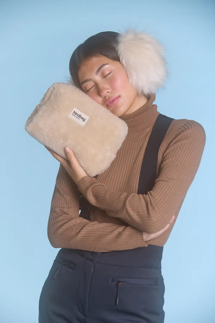Femme aux cheveux foncés portant un pull marron côtelé et un cache-oreilles en fausse fourrure beige, posant avec une trousse en fausse fourrure crème de la marque Hindbag contre sa joue. Fond bleu clair, ambiance douce et hivernale.