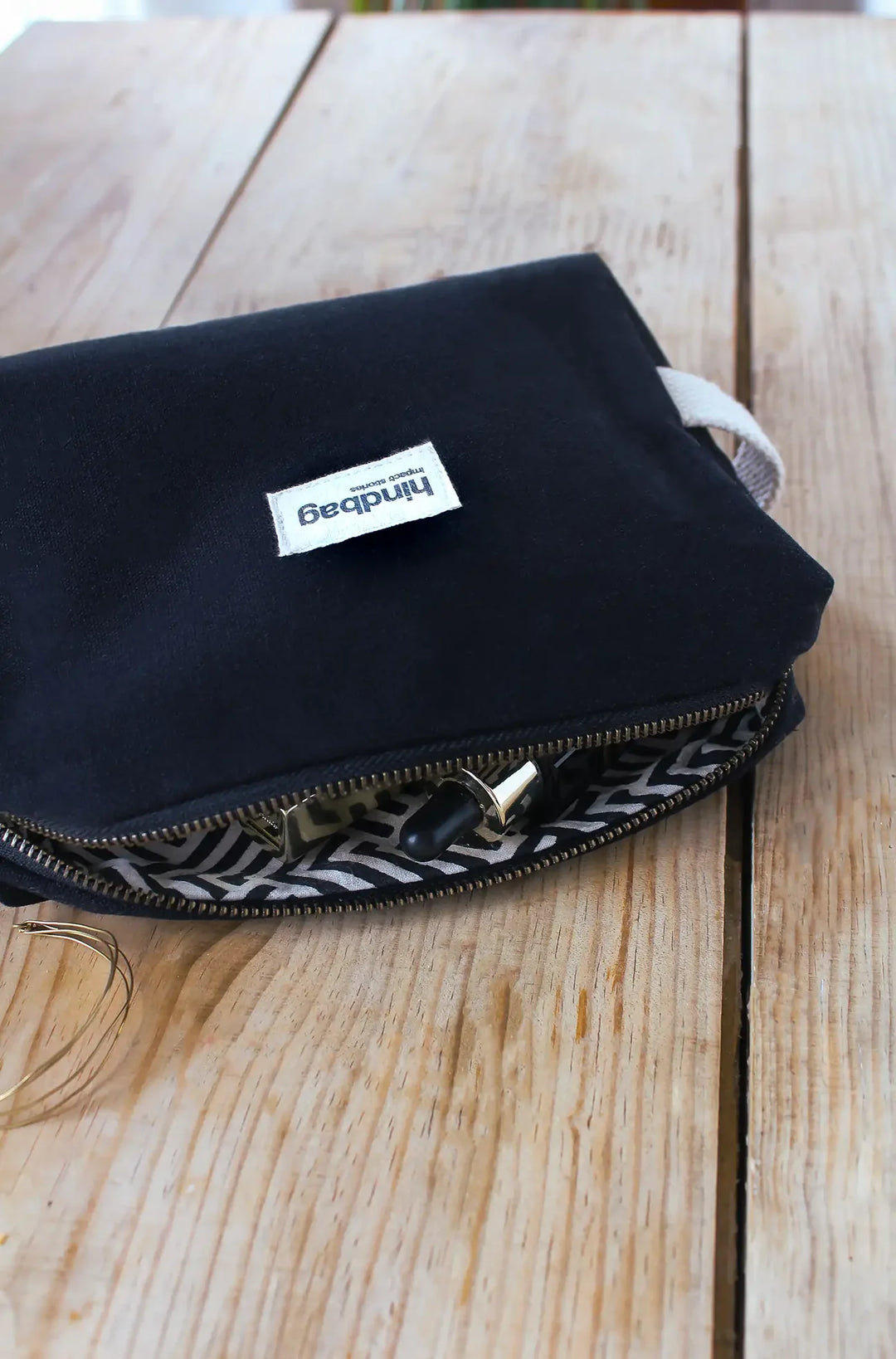 Trousse de toilette en toile de coton bio noir de la marque Hindbag, avec doublure intérieure imprimée graphique noir et blanc, posée sur une table en bois. La trousse est entrouverte, laissant apparaître des accessoires de beauté, dont un flacon compte-gouttes noir et doré. À côté, un bijou doré délicatement posé sur la table.