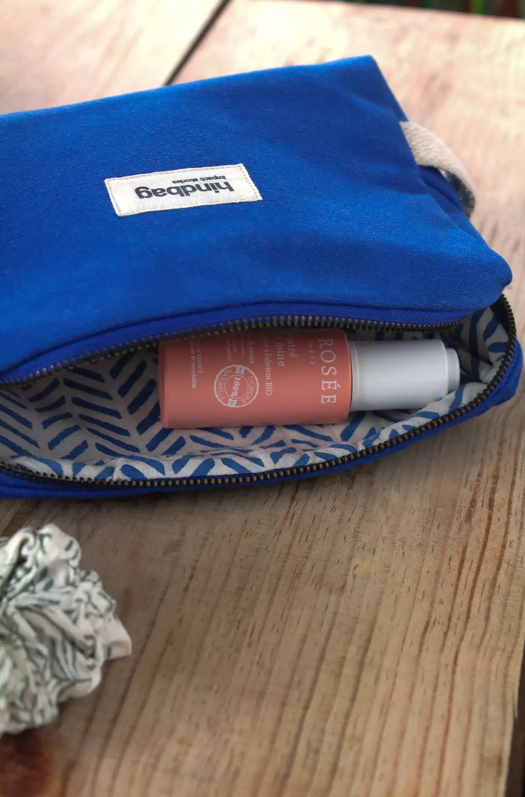 Trousse de toilette en toile de coton bio bleu électrique de la marque Hindbag, avec doublure intérieure imprimée feuillage bleu et blanc, posée sur une table en bois. À l'intérieur, un flacon rose de la marque La Rosée, et à côté, un chouchou en tissu à motif végétal.