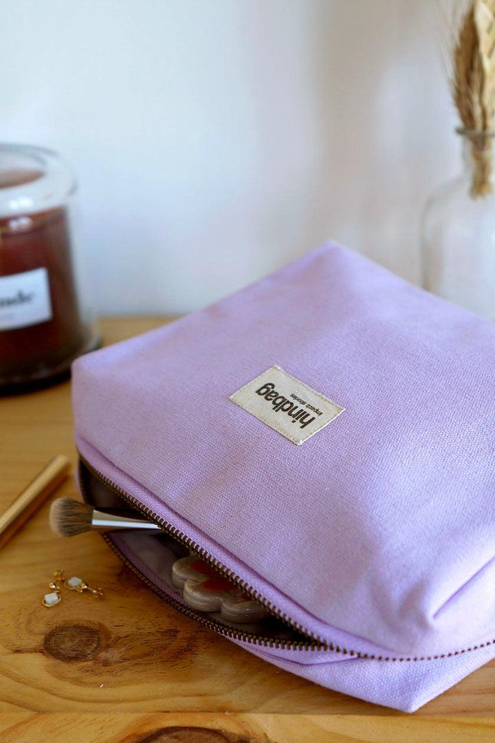 Trousse de toilette en toile de coton bio couleur lilas de la marque Hindbag, posée sur une table en bois. La trousse est légèrement ouverte, laissant apparaître un pinceau de maquillage, une palette de fards et des accessoires beauté. À côté, une bougie parfumée, un crayon doré et une paire de boucles d'oreilles minimalistes.