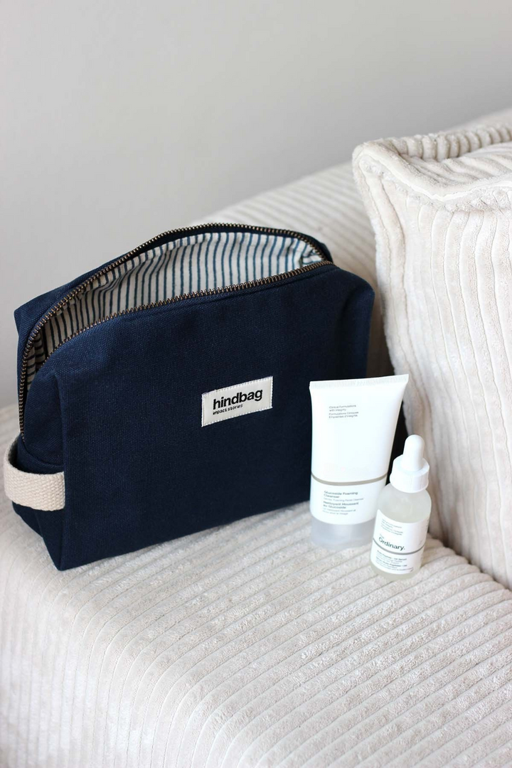Trousse de toilette en toile de coton bio bleu marine de la marque Hindbag, avec doublure intérieure rayée bleu et blanc, posée sur un canapé en velours côtelé crème. À côté, deux produits de soin visage au packaging minimaliste blanc.