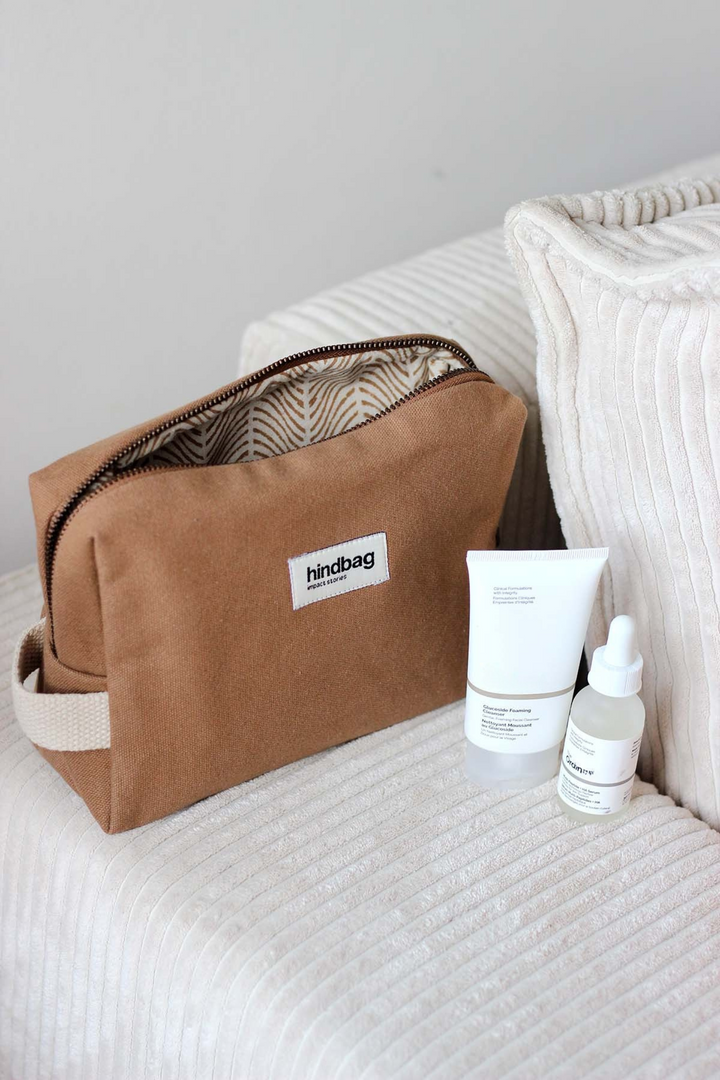 Trousse de toilette en toile de coton bio couleur camel de la marque Hindbag, avec doublure imprimée graphique beige et marron, posée sur un canapé en velours côtelé crème. À côté, deux produits de soin visage au packaging minimaliste blanc.