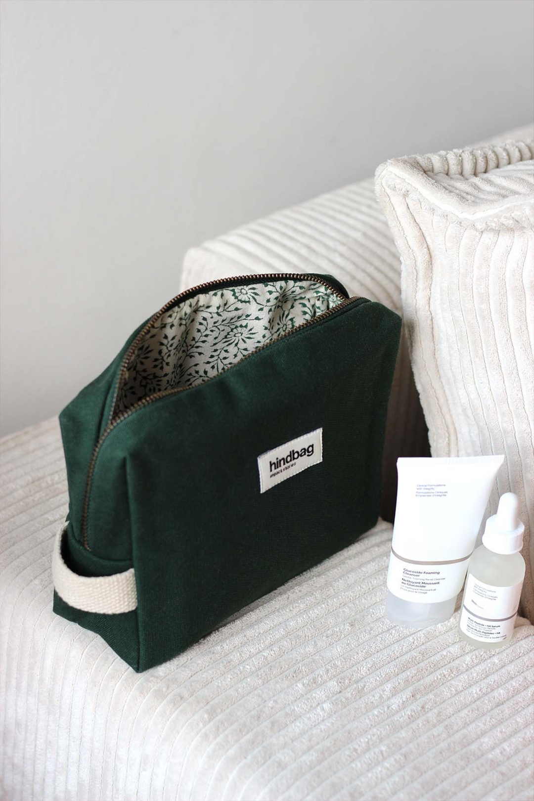 Trousse de toilette en toile de coton bio vert foncé de la marque Hindbag, avec doublure imprimée végétale, posée sur un canapé en velours côtelé crème. À côté, deux produits de soin visage en packaging minimaliste blanc.