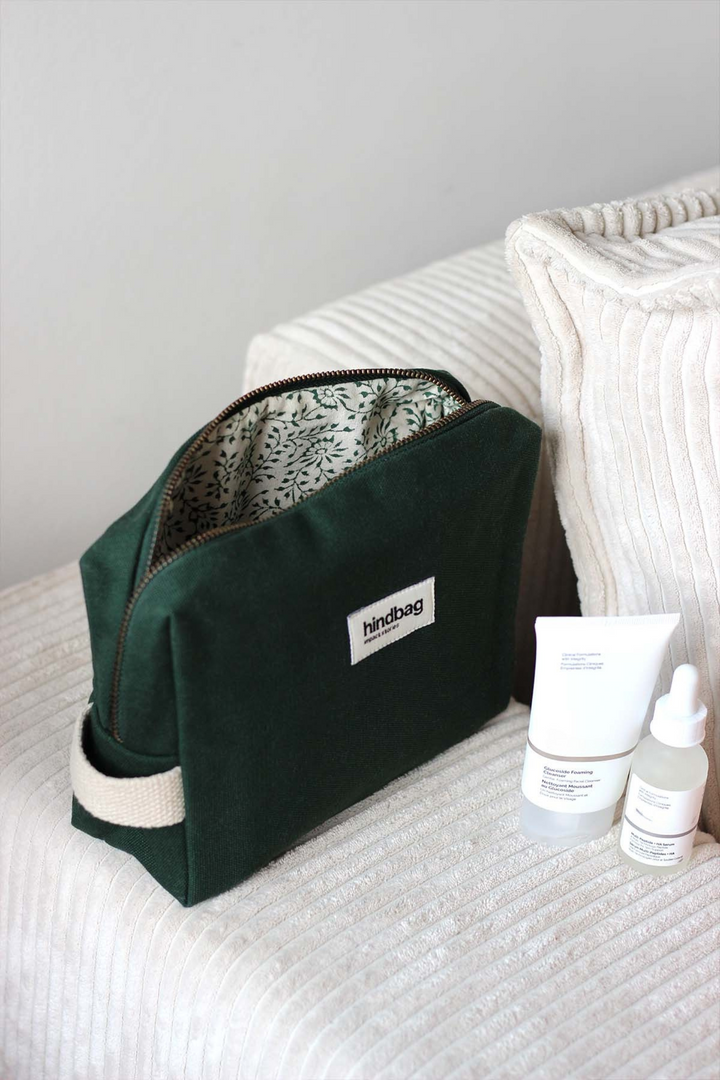 Trousse de toilette en toile de coton bio vert foncé de la marque Hindbag, avec doublure imprimée végétale, posée sur un canapé en velours côtelé crème. À côté, deux produits de soin visage en packaging minimaliste blanc.