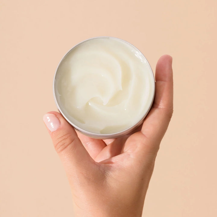 Les Petits Prödiges Baume Magique tenu en main, montrant la texture crémeuse du baume multi-usages 100% d'origine naturelle. Pot ouvert, main avec ongles manucurés, sur fond beige.