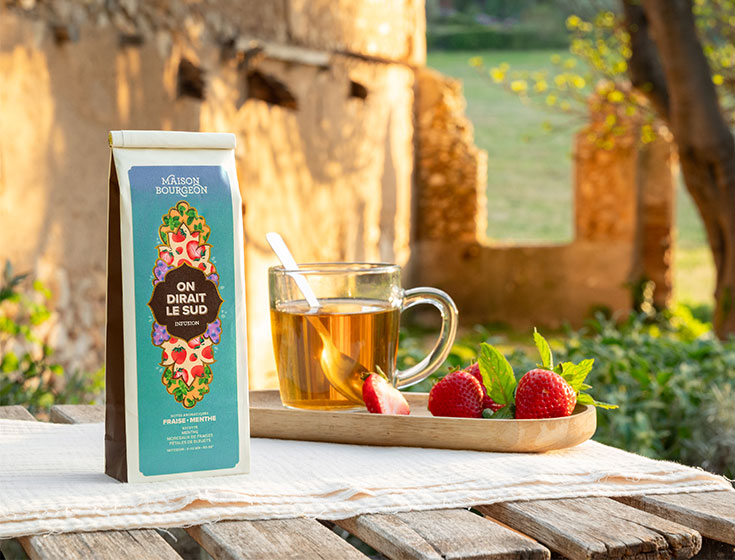 Maison Bourgeon On Dirait Le Sud infusion posée sur une table en bois en extérieur, accompagnée d'un verre de thé infusé, de fraises fraîches et de feuilles de menthe, avec un paysage rural en arrière-plan.