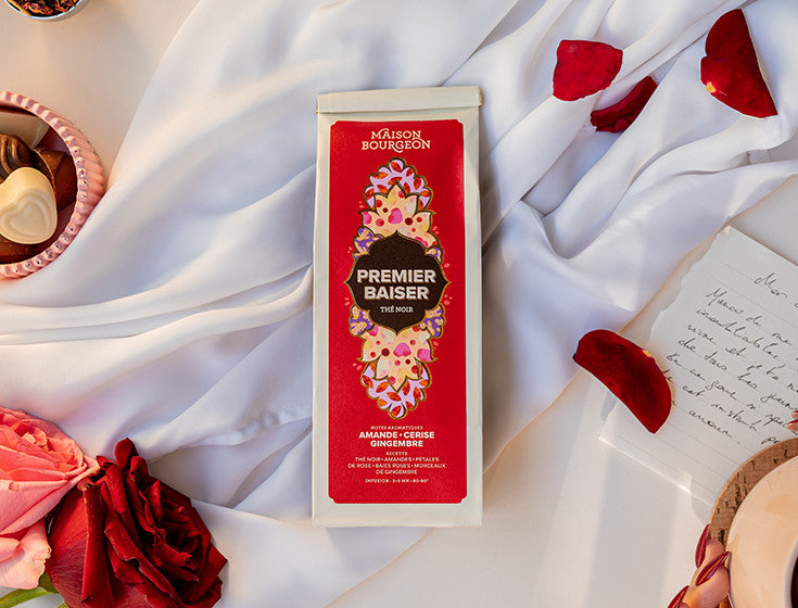 Paquet de thé noir Maison Bourgeon Premier Baiser posé sur un tissu blanc avec des pétales de rose et des roses rouges à côté.