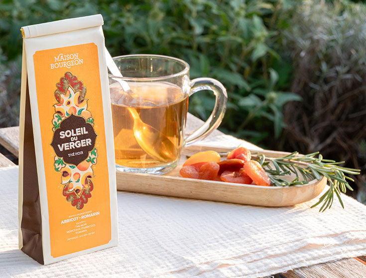 Paquet de thé noir Maison Bourgeon Soleil du Verger avec une tasse de thé, des morceaux d'abricots secs et du romarin en arrière-plan.