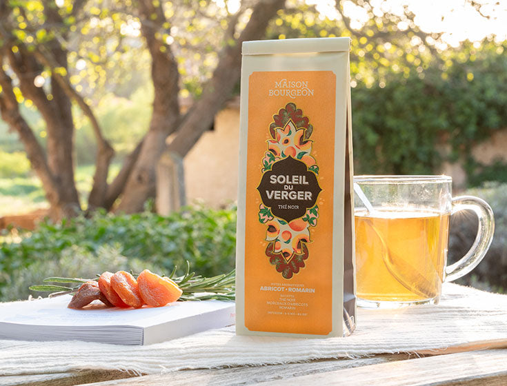 Paquet de thé noir Maison Bourgeon Soleil du Verger avec une tasse de thé et des abricots secs sur une table en extérieur, avec des arbres en arrière-plan.