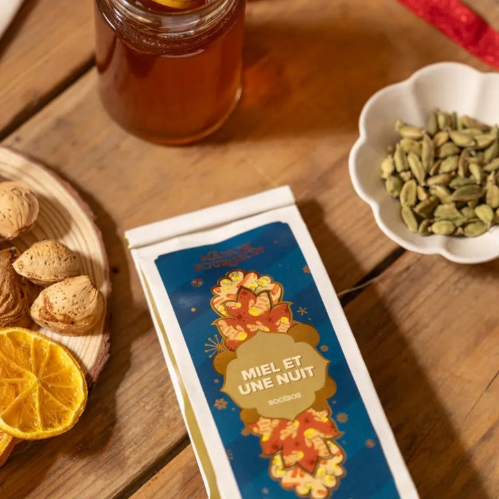 Rooibos Miel et une Nuit Maison Bourgeon posé sur une table avec cardamome, orange séchée et amandes